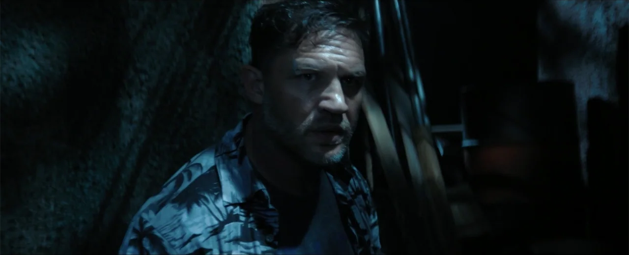 Venom: The Last Dance (2024) movie still: closeup — A dimly lit, narrow interior space features rough textured walls and wooden supports. A man…