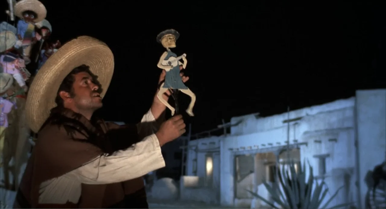 The Spikes Gang (1974) movie still: medium — An outdoor market at night features a man in a sombrero holding a puppet. The man inspects a small…