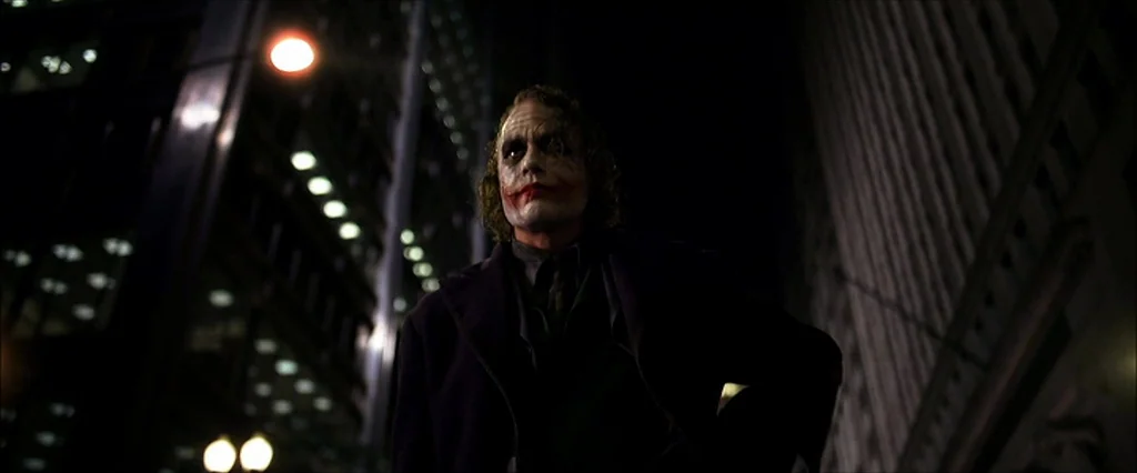 The Dark Knight (2008) movie still: medium — A dark, narrow urban street is lined with tall, indistinct skyscrapers at night. The Joker stands in…