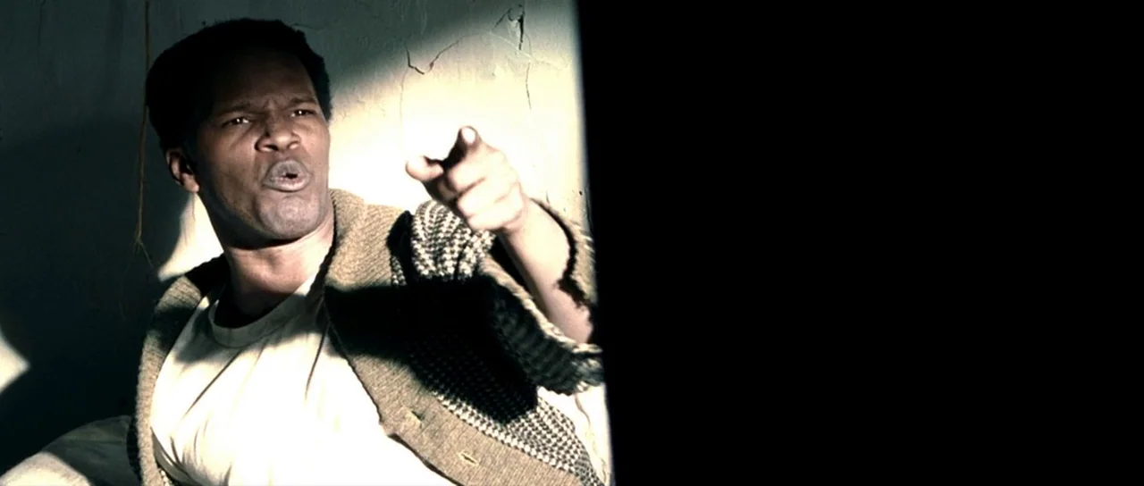 Ali (2001) movie still: medium — A dimly lit room features a textured white wall with visible cracks and shadows. A man points his finger toward the…