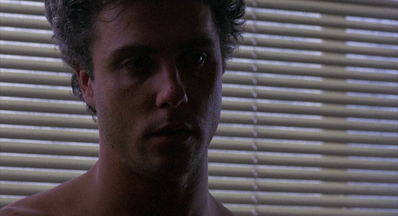 To Live and Die in L.A. (1985) movie still: closeup — A man stands before a window covered by horizontal blinds at night. He remains shirtless and…