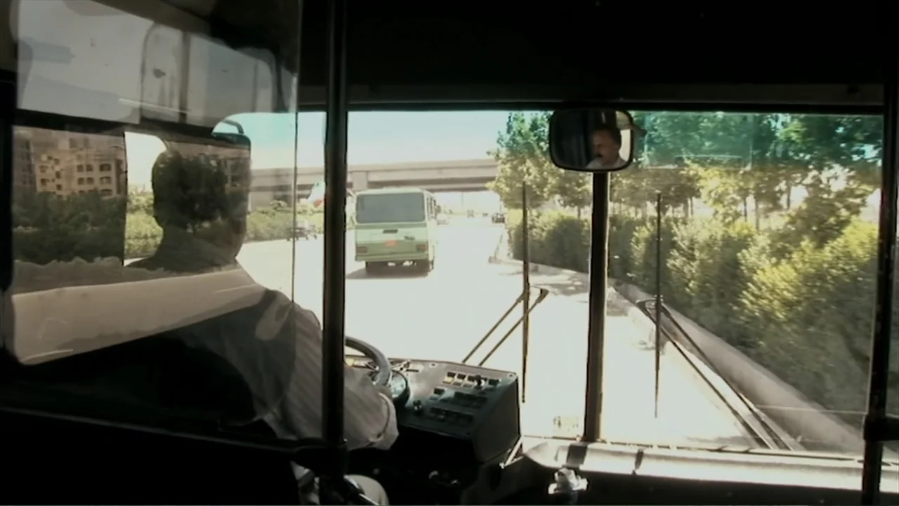 Offside (2015) movie still: medium — The view from inside a bus cabin shows the driver's back and the road ahead. A green minibus travels along a…