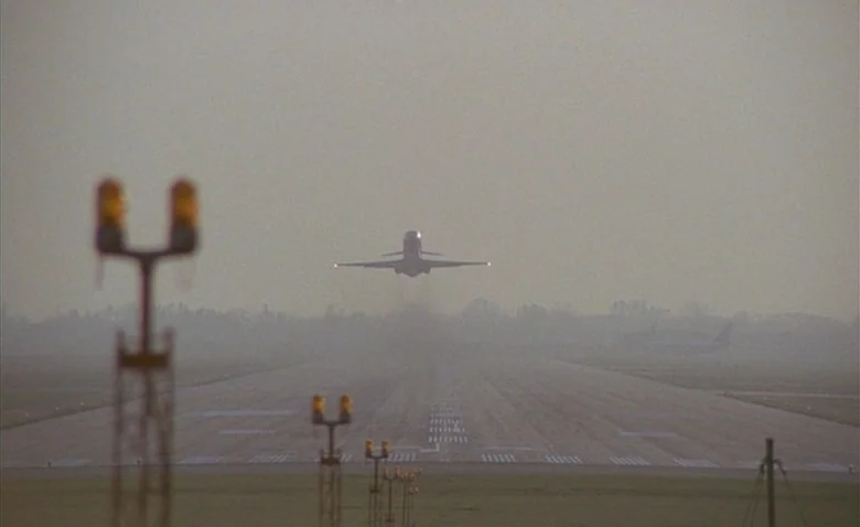 Sweet Movie (1974) movie still: wide — An asphalt runway stretches into a hazy, fog-filled horizon under a bright sky. A passenger jet airplane…
