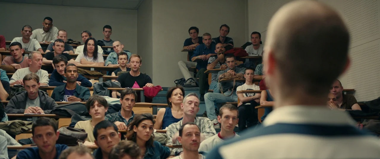 BPM (Beats Per Minute) (2017) movie still: medium — A tiered lecture hall is filled with rows of students sitting at desks. The view is from behind…