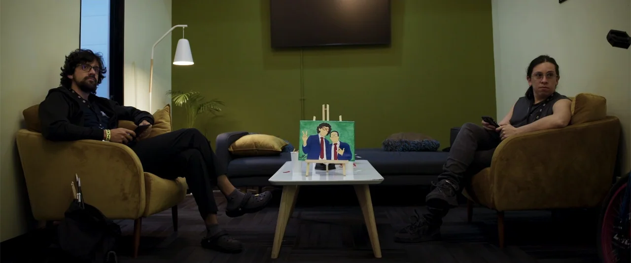 Making of: F*CKS NEWS (2025) movie still: medium — A modern waiting room features olive green walls and two yellow armchairs facing inward. Two men…