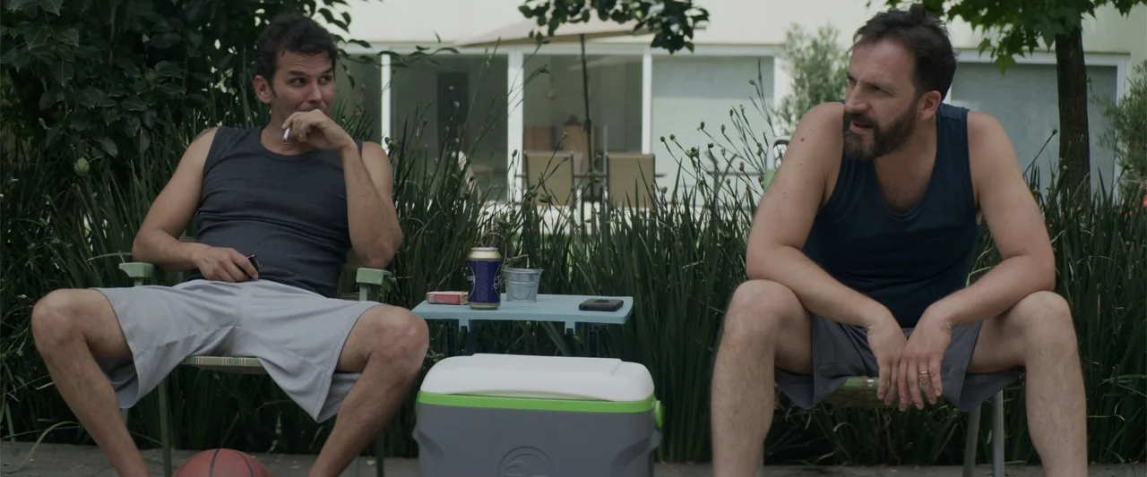 Human Animals (2020) movie still: medium — Two men sit in folding chairs on a residential patio next to tall grass. They are having a conversation…