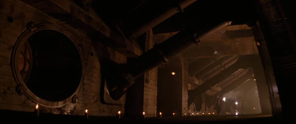 Alien 3 (1992) movie still: wide — A dark subterranean concrete hallway features large circular bulkhead portals on the wall. Rows of lit candles…