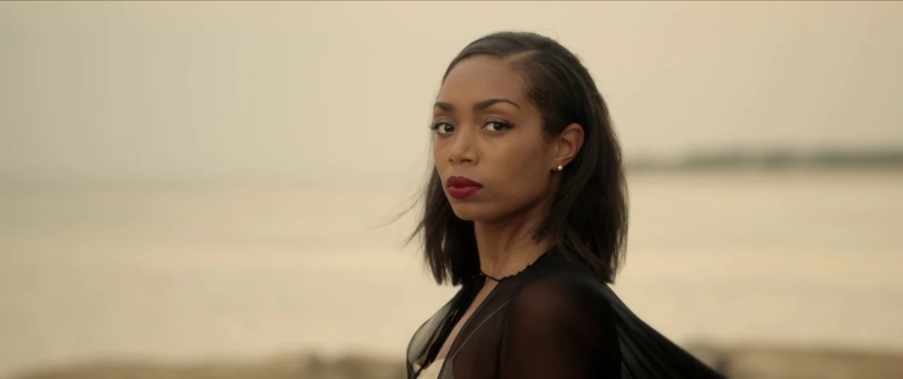 Da Sweet Blood of Jesus (2014) movie still: closeup — A woman stands before a large body of water under an overcast sky. She turns her head to look…