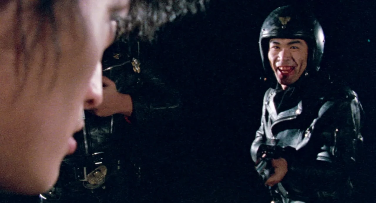 Burst City (1982) movie still: medium — A dark outdoor setting at night features a man wearing a motorcycle helmet. He stares toward an unseen…