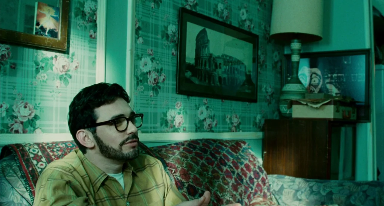 Howl (2010) movie still: medium — A man sits on a couch in front of walls covered in floral wallpaper. He gestures with his hands while looking…