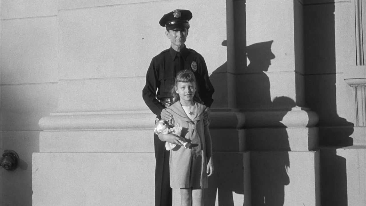 The Naked Kiss (1964) movie still: fullbody — A smooth stone wall of a building facade serves as a backdrop. A police officer stands behind a young…