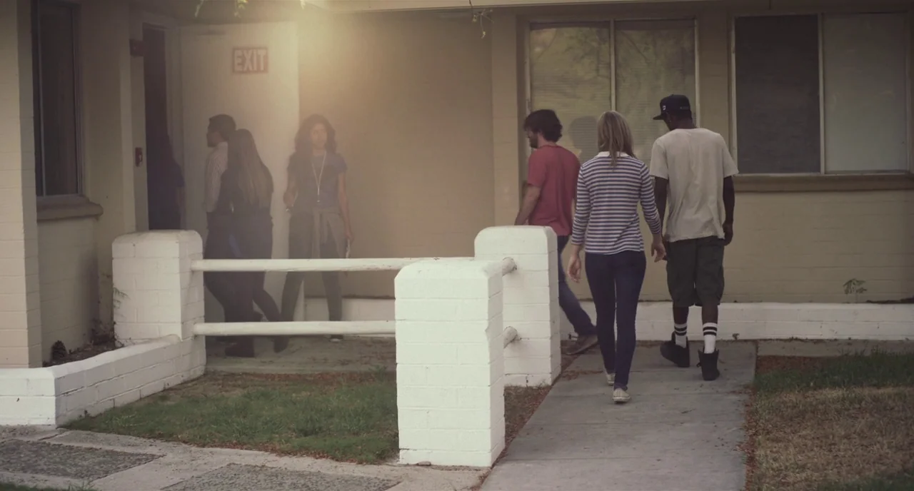 Short Term 12 (2013) movie still: wide — A group of young people walks along a concrete walkway past a beige building. Several individuals approach…