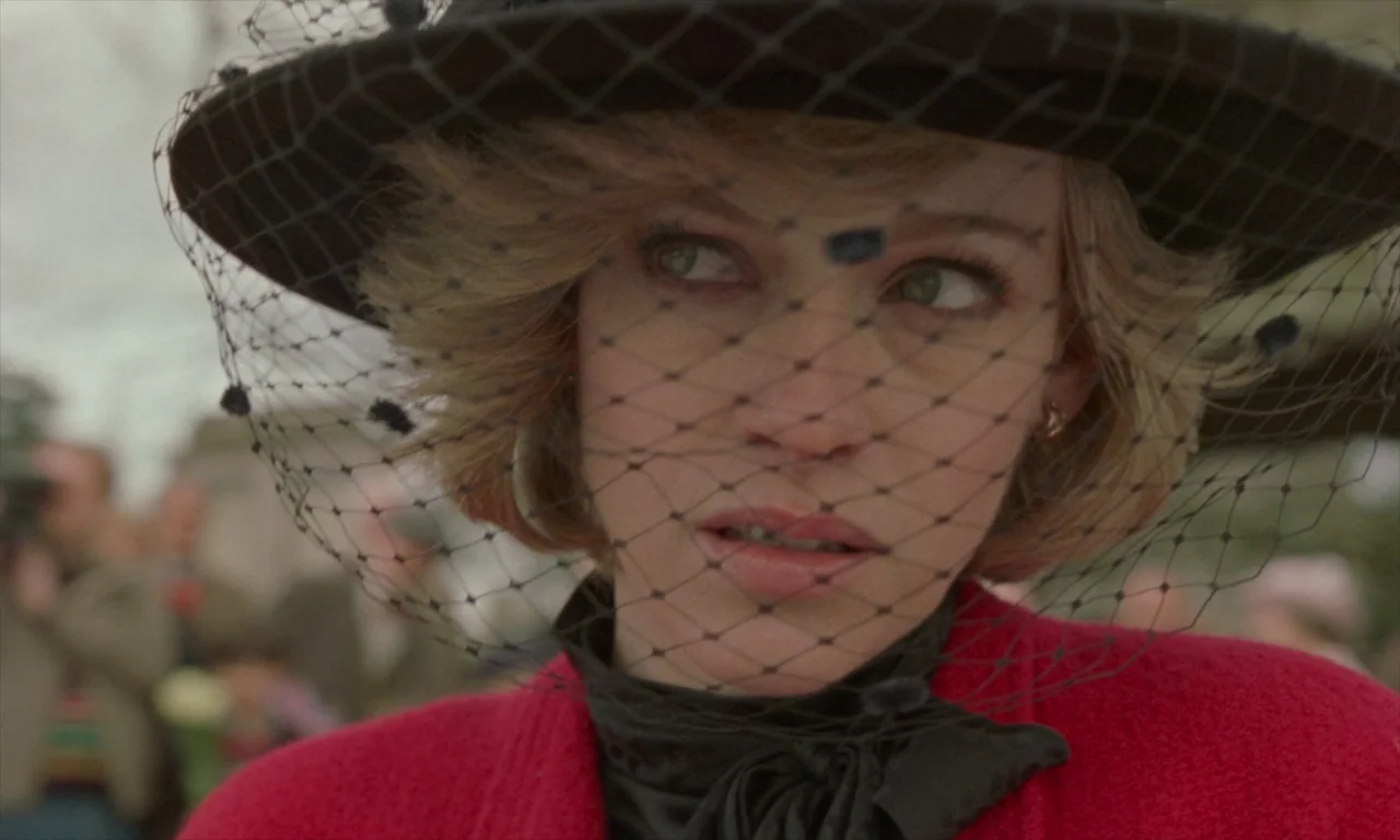 Spencer (2021) movie still: closeup — A woman wears a black hat with a veil and a red coat outdoors. She looks toward the side while surrounded by a…
