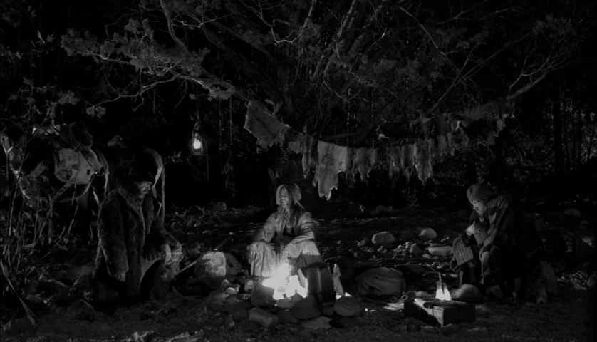 Dead Man (1995) movie still: wide — A dark, wooded clearing at night features a small campfire between three seated figures. Animal pelts hang…