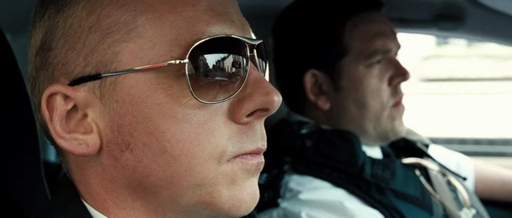 Hot Fuzz (2007) movie still: closeup — A view inside a moving police car showing two uniformed officers in the front. The foreground officer wears…