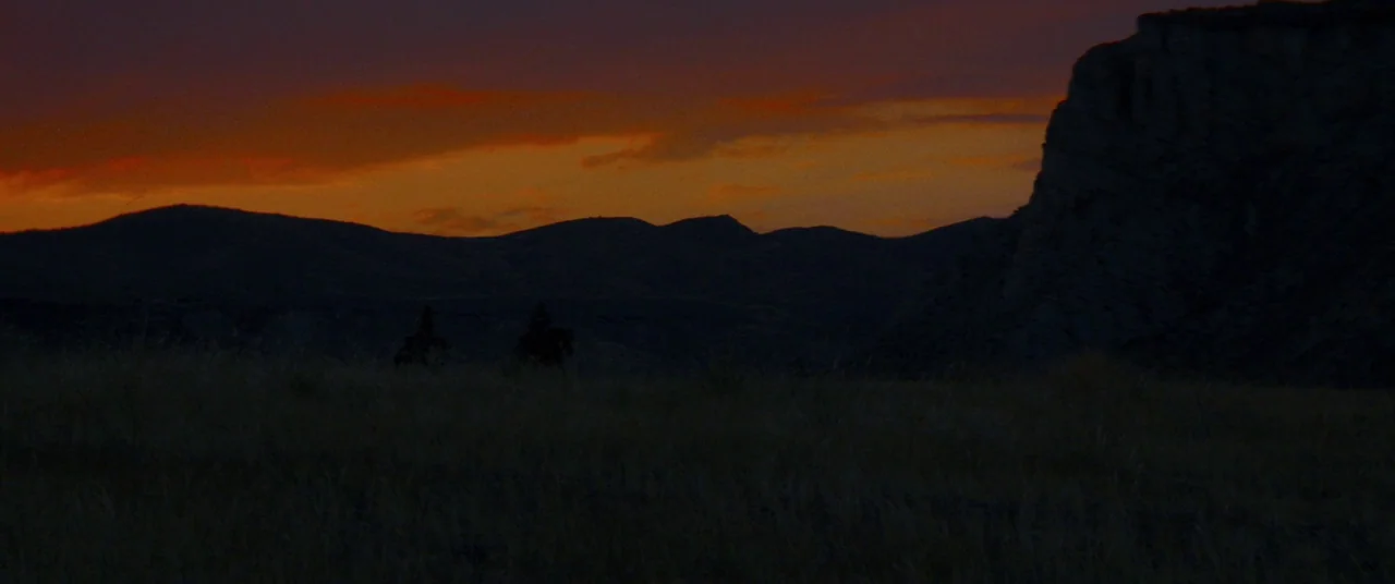The Sisters Brothers (2018) movie still: wide — A vast, dark, grassy plain extends toward distant rolling hills under a sunset sky. Two riders on…