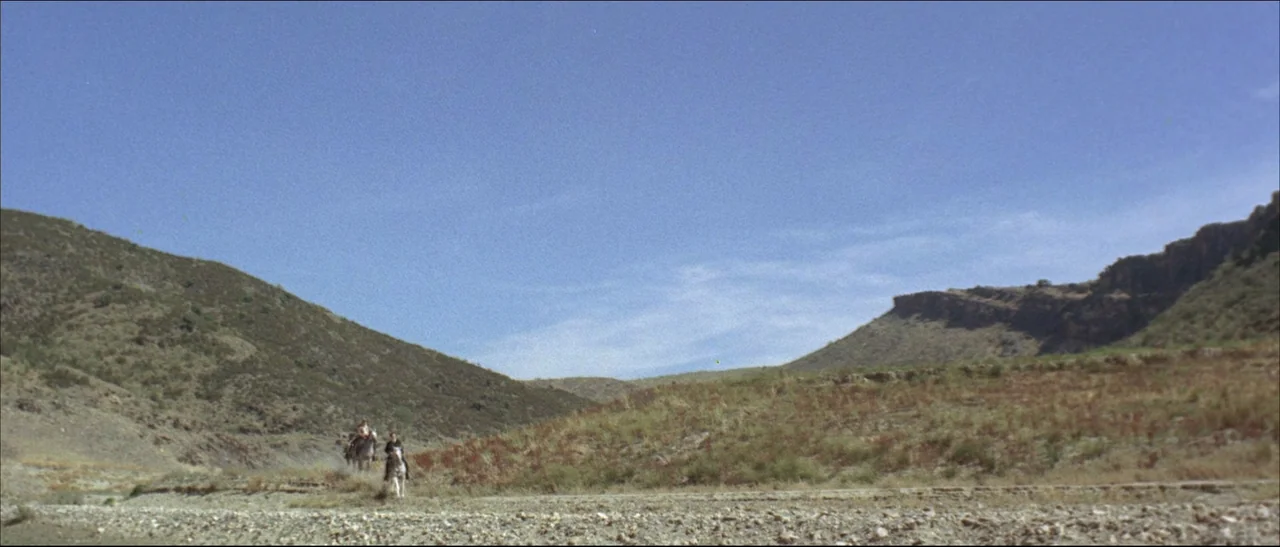 The Mercenary (1968) movie still: wide — A wide canyon landscape with rocky terrain, sparse dry vegetation, and a cloudless sky. Two figures riding…