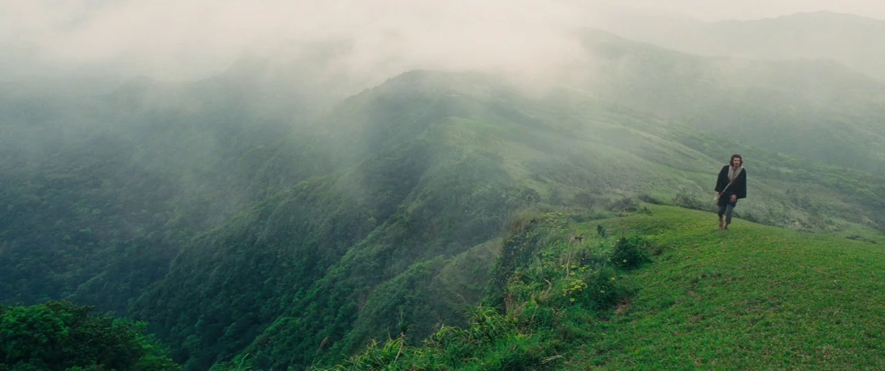 Silence (2016) movie still: wide — A green mountain ridge stretches into the distance under heavy, low-hanging clouds and fog. A single man walks…