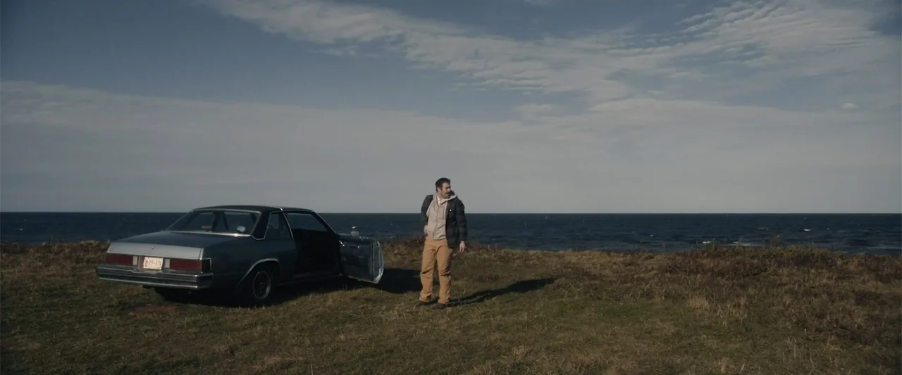 A Small Fortune (2021) movie still: fullbody — A grassy coastal ridge borders a dark, choppy ocean under an overcast sky. A man in casual clothing…
