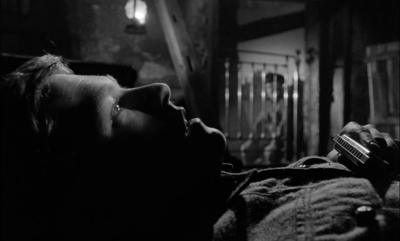 King & Country (1964) movie still: closeup — An attic space with wooden beams and a metal cot visible in the background. A man lies on his back…