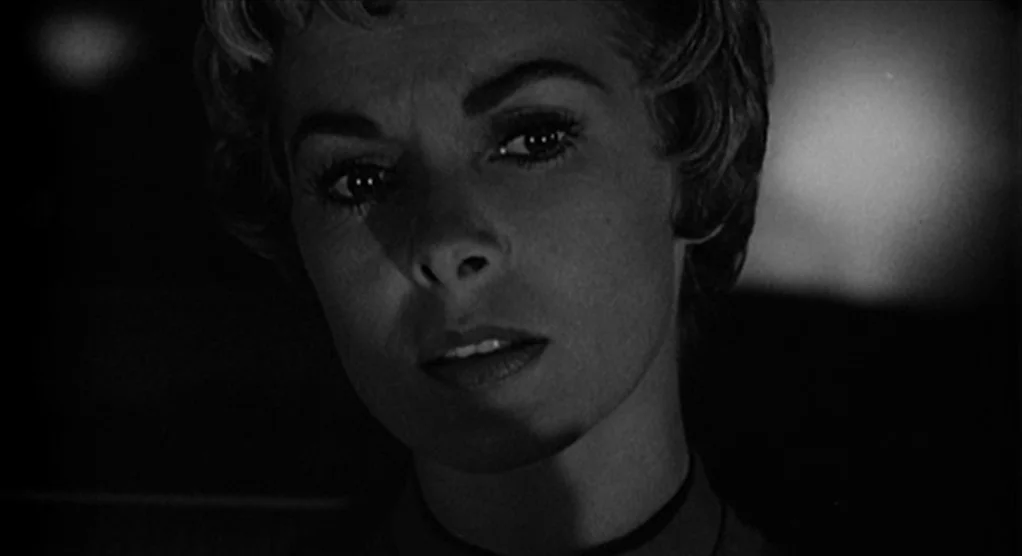 Psycho movie still: closeup — A dimly lit car interior at night with a dashboard reflecting muted light. A woman stares straight ahead with a set…