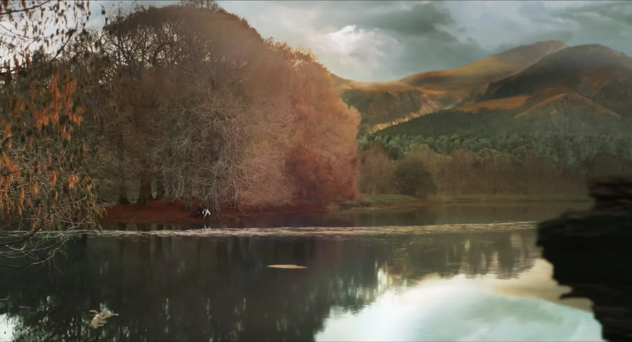 Echoes Of A Crime (2025) movie still: wide — A tranquil lake is framed by dense, orange-hued autumn trees and distant rolling mountains. A solitary…