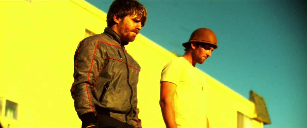 Bellflower (2011) movie still: medium — A bright yellow wall creates a backdrop for two men standing in sunlight. The men look toward the ground…