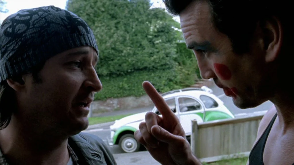 Dead Man’s Shoes (2004) movie still: medium — A suburban street scene with a white and green vintage car in the background. Two men engage in a…