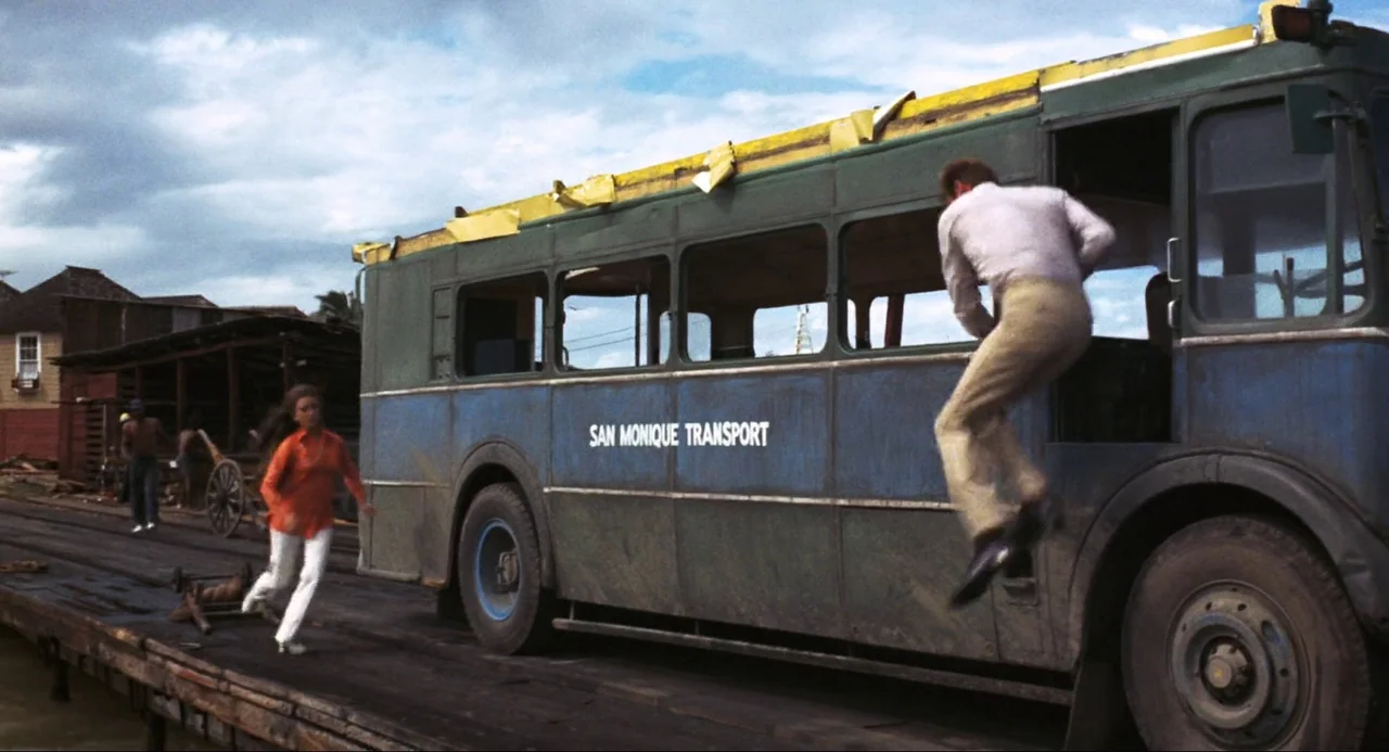 Live and Let Die (1973) movie still: fullbody — A weathered bus labeled San Monique Transport sits on a wooden dock exterior. A man jumps into the…
