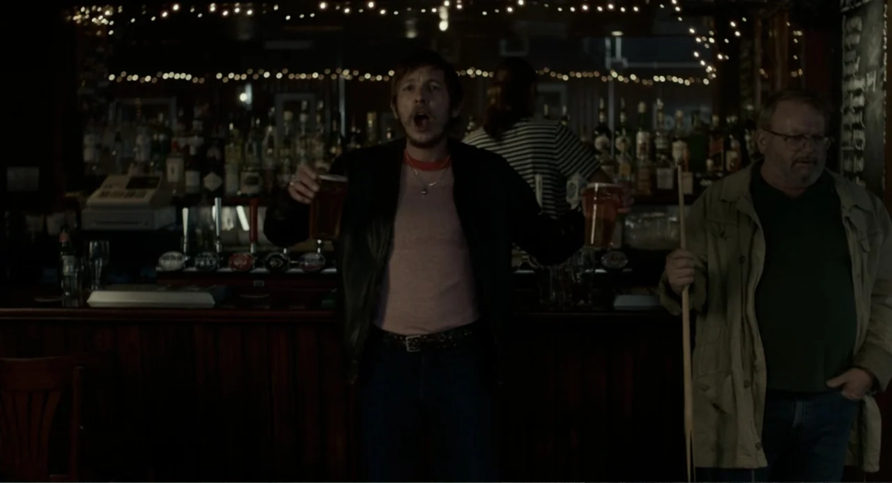 Back to Black (2024) movie still: medium — A dimly lit pub with a wooden counter and rows of liquor bottles behind. A man in a leather jacket stands…