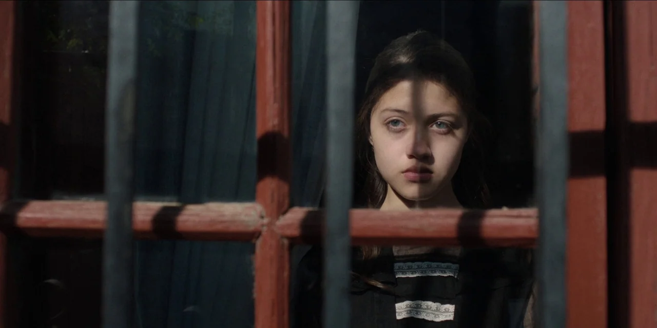 Pedro Páramo (2024) movie still: closeup — A young girl stands behind a wooden window frame with dark vertical metal bars. She looks forward with…