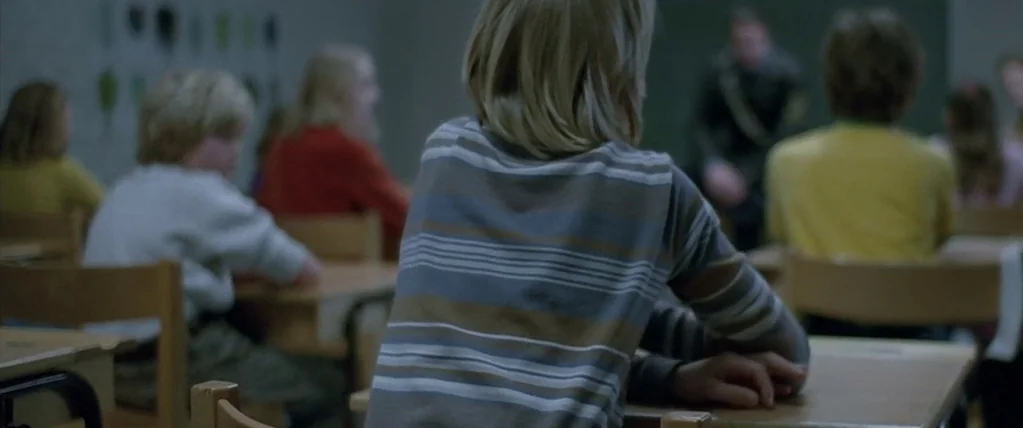 Let The Right One In (2008) movie still: medium — A school classroom with students seated at wooden desks facing toward the front. The room has…