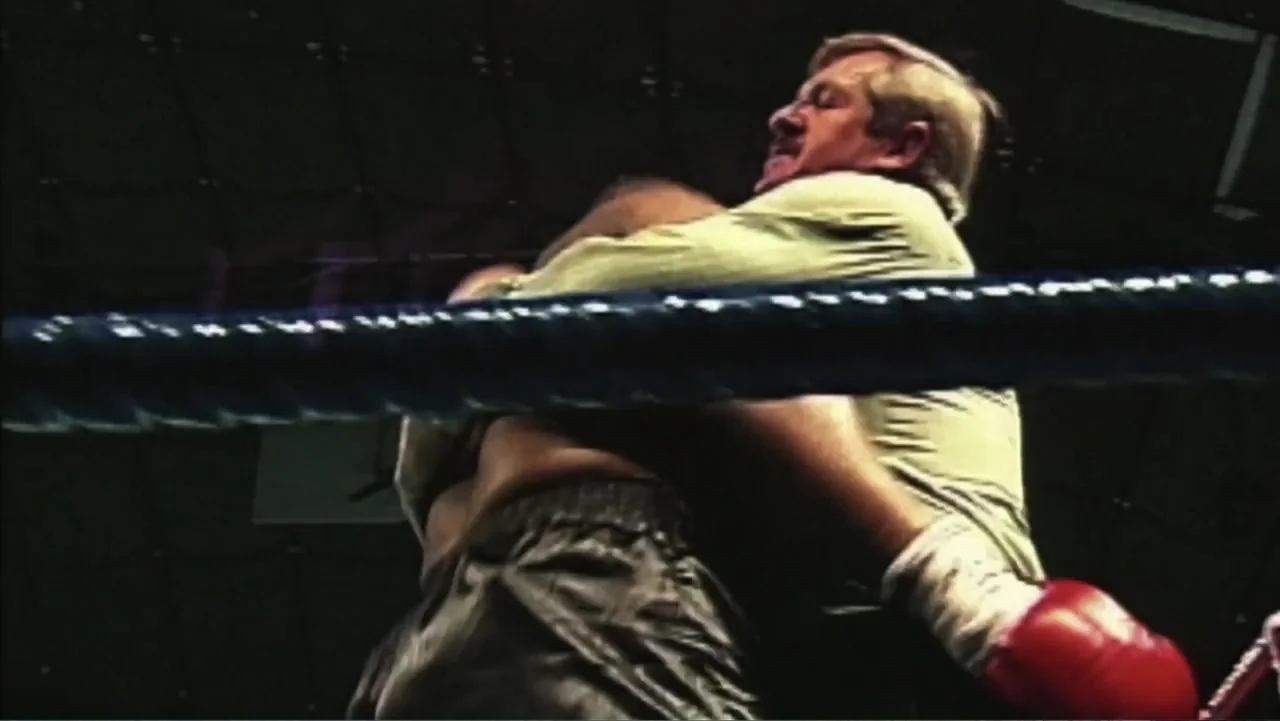 Mike Tyson: Undisputed Truth (2013) movie still: medium — A dimly lit boxing ring surrounded by dark ropes at night. Two men are engaged in a close…