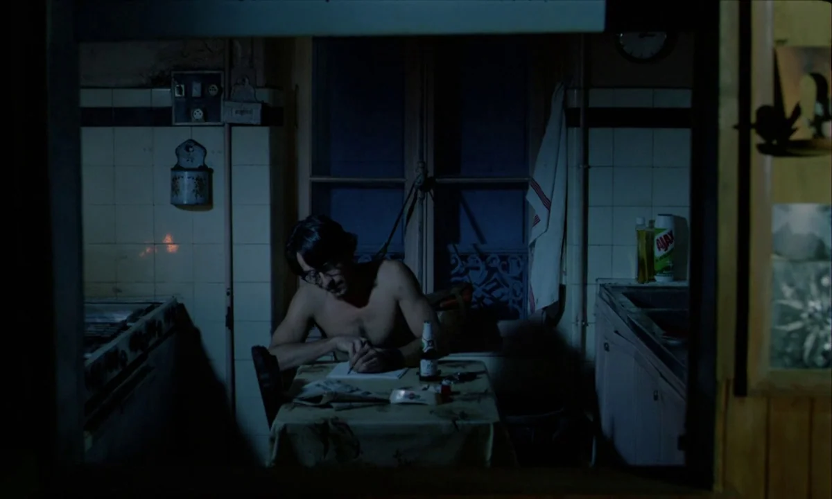 Betty Blue (1986) movie still: medium — A dimly lit 1970s kitchen features tiled walls, a small table, and household clutter. A shirtless man sits…