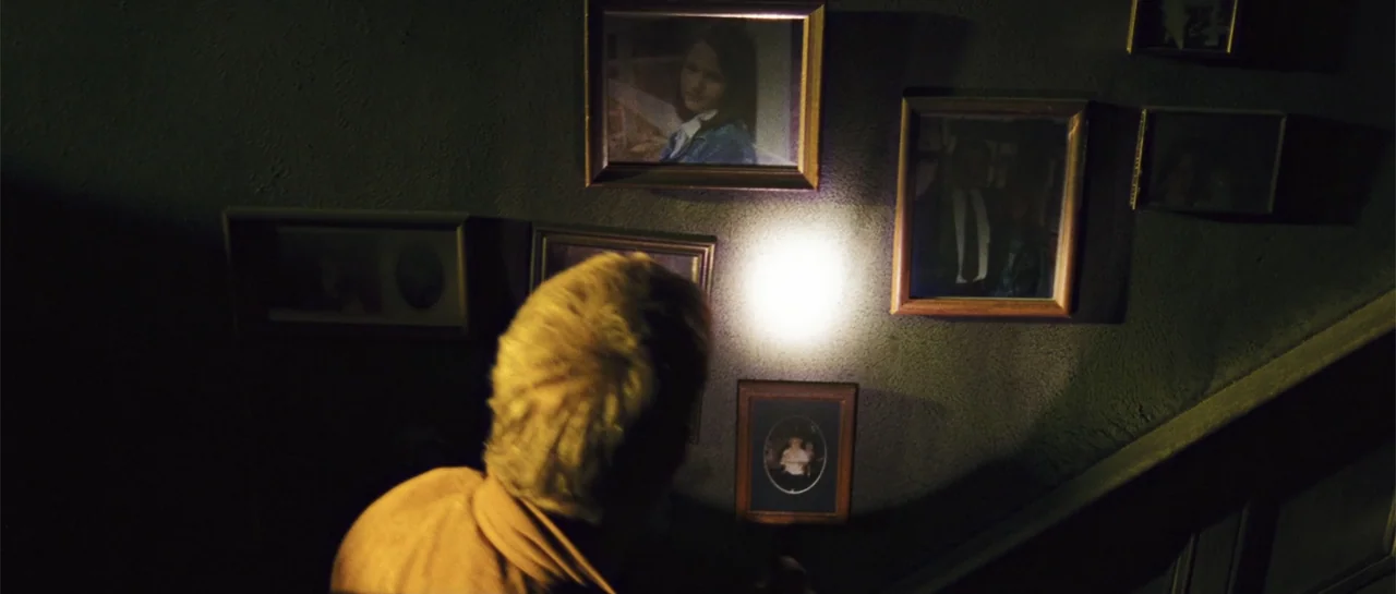 The New Daughter (2009) movie still: medium — A dimly lit staircase wall is covered with multiple framed vintage photographs. A person seen from…