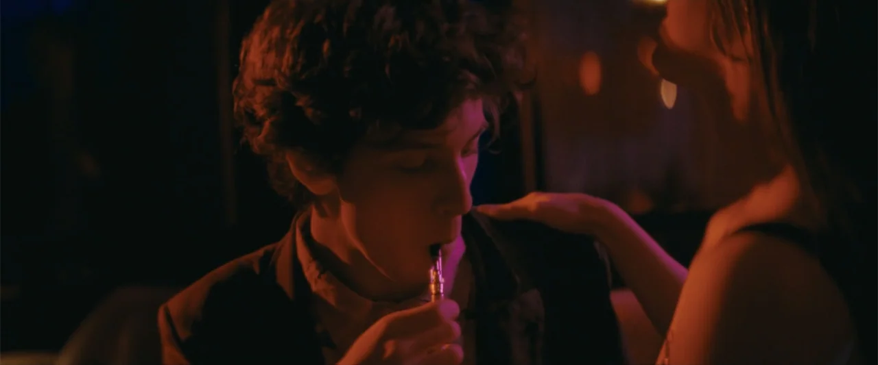 Anora (2024) movie still: closeup — A dark indoor space is bathed in intense red and purple neon light. A young man holds a small glowing vaping…