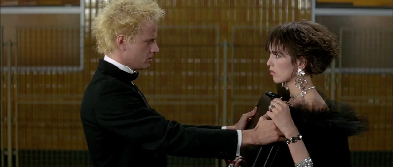 Subway (1985) movie still: medium — A locker room with rows of orange-brown lockers in the background behind a fence. A blonde man in a tuxedo…