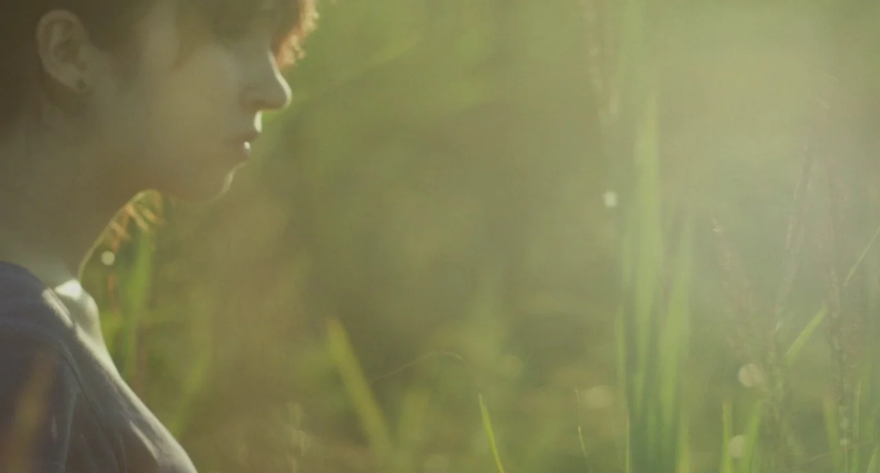 It Felt Like Love (2013) movie still: closeup — A woman stands in a field of tall grass during late afternoon light. She looks toward the side of…