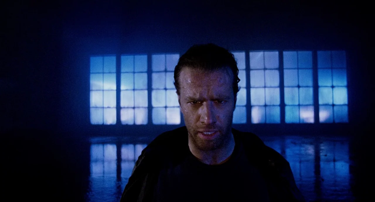 Highlander (1986) movie still: medium — A dark industrial space features large glowing blue window panes against a black background. A sweaty man…