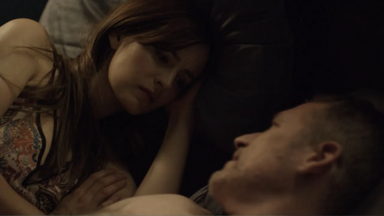 Totem (2022) movie still: closeup — A dimly lit bedroom shows two people lying in bed together in low light. The woman rests her head near the man…