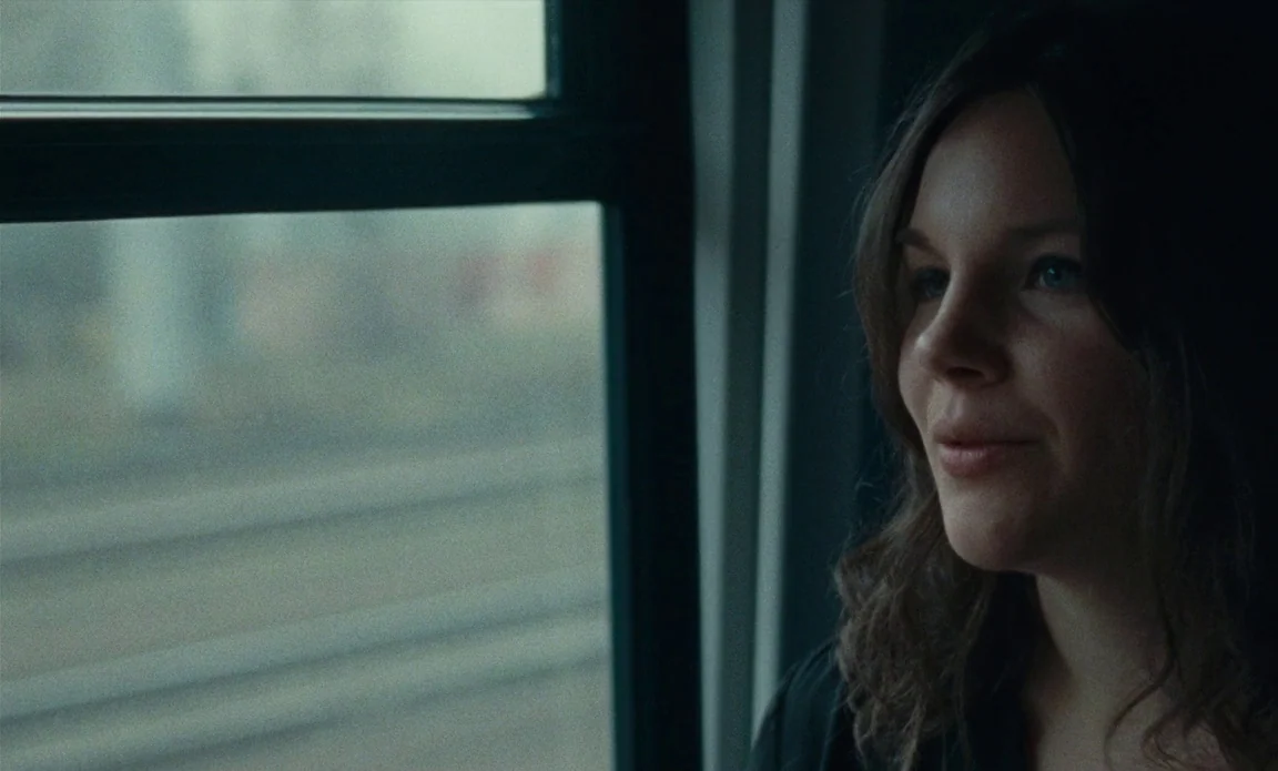 High Life (2018) movie still: closeup — A woman looks out the window of a moving train during the day. Blurry, out-of-focus train tracks are visible…
