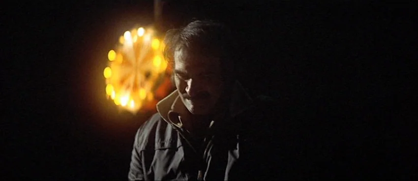 California Split (1974) movie still: medium — A man stands in darkness against a backdrop of a blurred, glowing fairground attraction. He is looking…