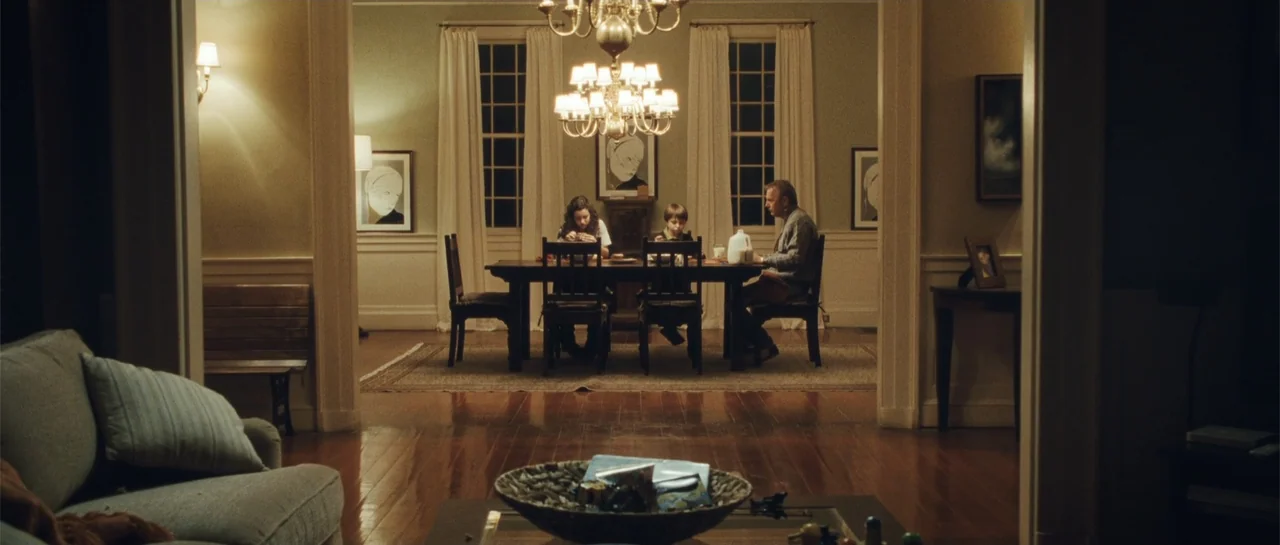 The New Daughter (2009) movie still: wide — A residential home with a view from the living room into the dining area. A mother, son, and father sit…