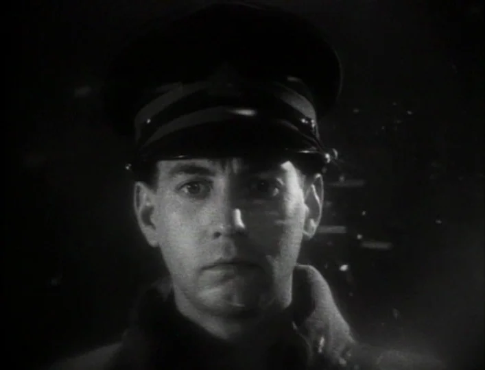 Archangel (1990) movie still: closeup — A man in a military uniform is centered against a pitch black background. The subject maintains a frontal…
