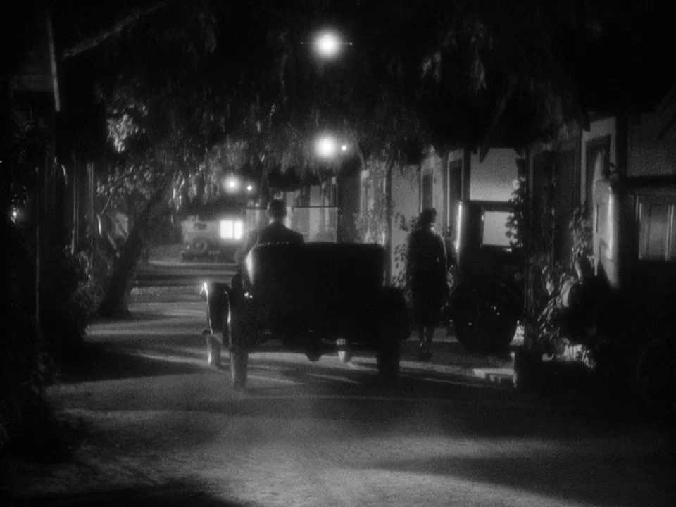 It Happened One Night (1934) movie still: wide — A quiet residential street at night is illuminated by dim, high-mounted overhead lights. A person…