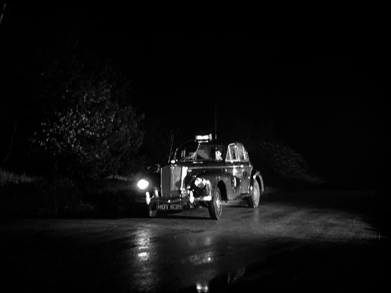 The Sleeping Tiger (1954) movie still: wide — A dark, empty road at night is illuminated by the headlights of a car. A vintage police car travels…