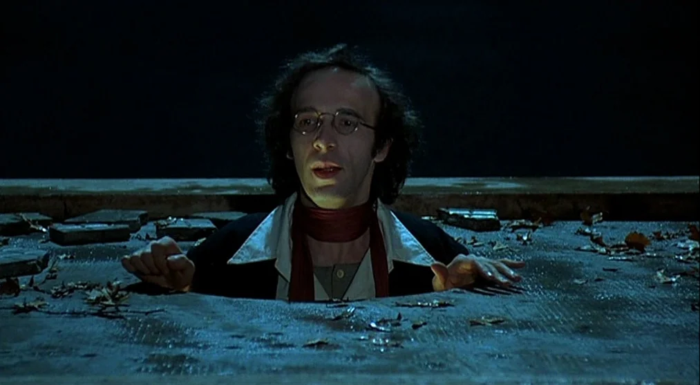 The Voice of the Moon (1990) movie still: medium — A man emerges from a rectangular opening in a dark, paved surface. Dried leaves and debris litter…