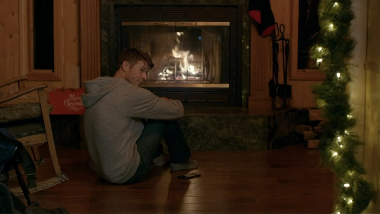 Alex & Emma (2003) movie still: medium — A rustic living room with wooden walls and a central glass-enclosed stone fireplace. A young man wearing a…