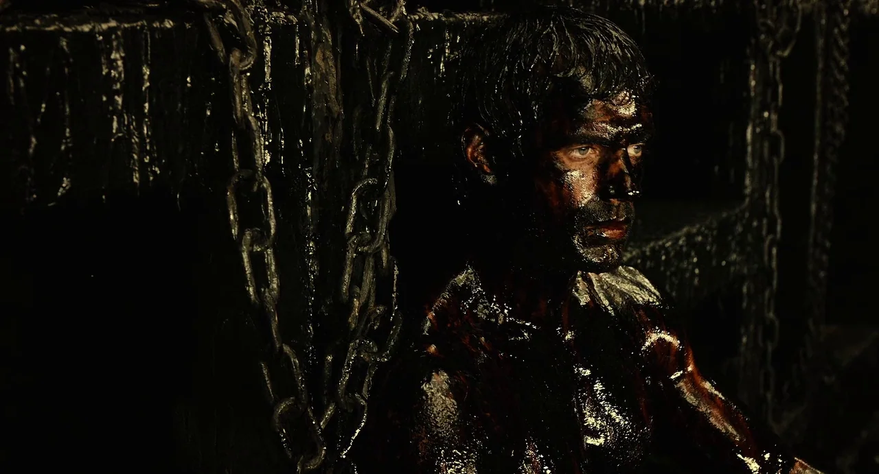 The Immortals (2011) movie still: medium — A dark, cramped space features heavy iron chains hanging against a textured black wall. A shirtless man…