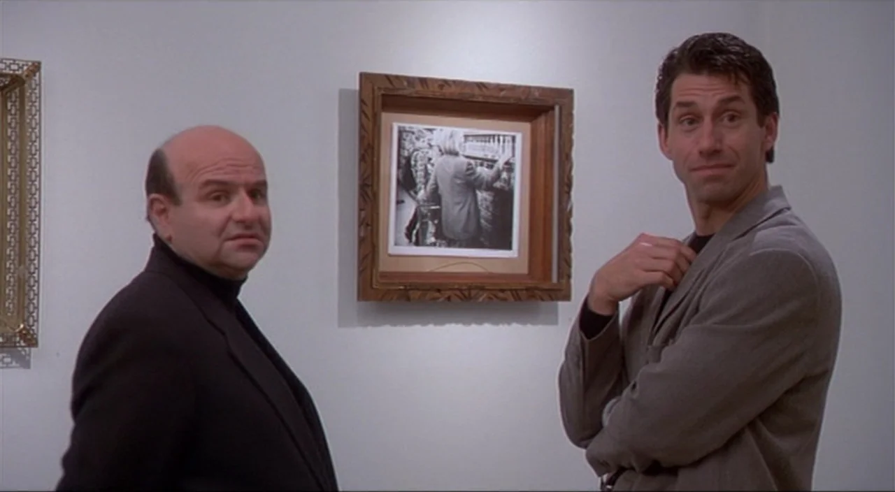 Pecker movie still: medium — Two men stand in front of a white wall decorated with framed art pieces. They look toward the camera while situated in…